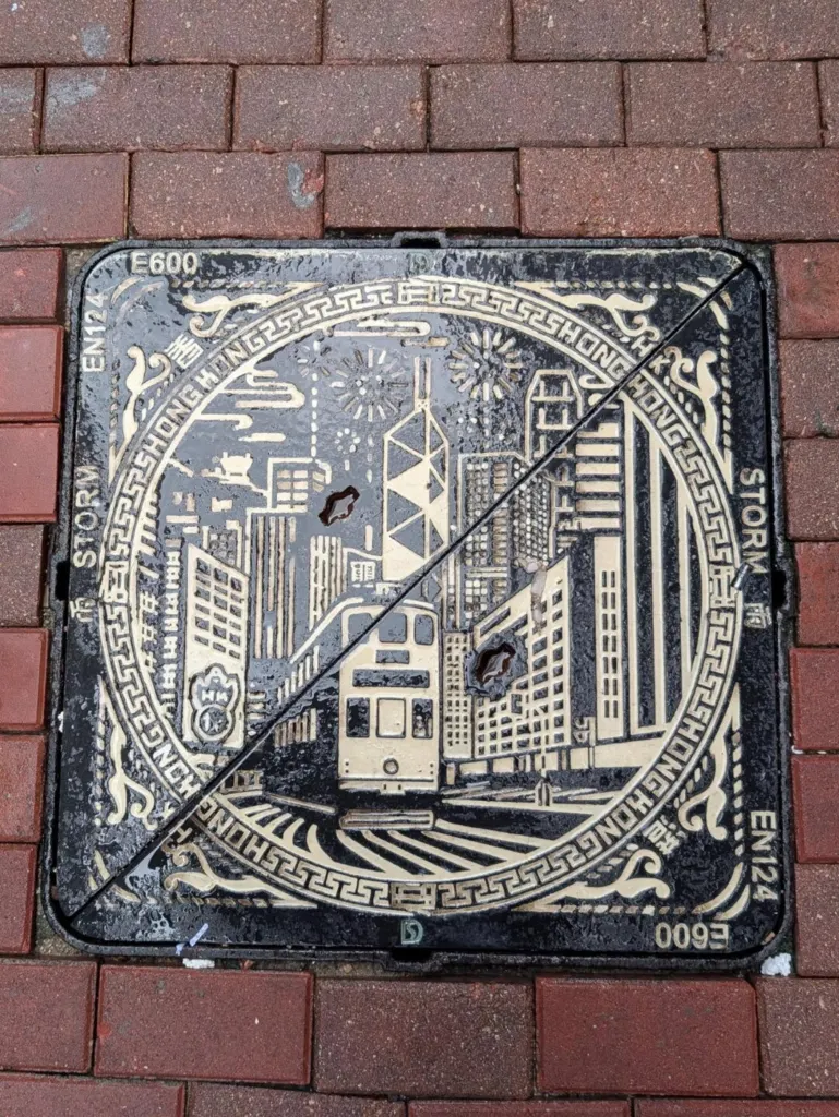 A Hong Kong manhole cover cast with a tram, skyline and harbour, set in red brick paving