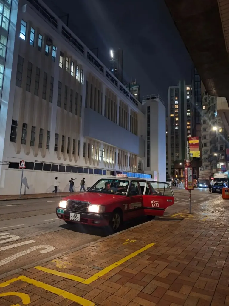 A red Hong Kong taxi on a wet street at night, below a tall grid-faced building